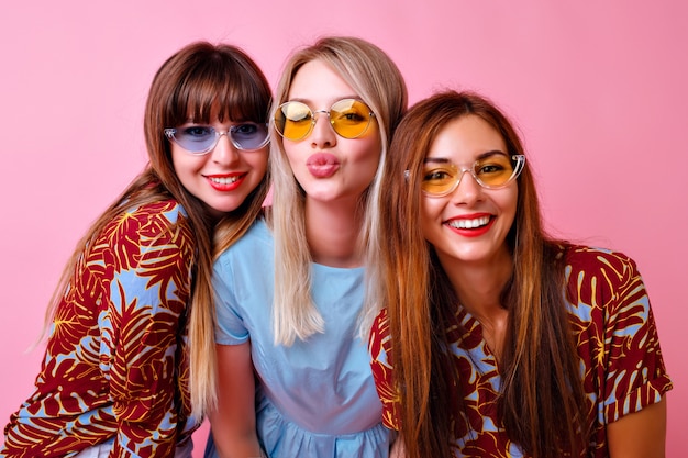 Adorable grupo de chicas con estilo sonriendo y enviando besos, ropa con estampado tropical súper moderno y gafas de colores estilo años 90, mejores amigas disfrutan del tiempo juntas, pared rosa