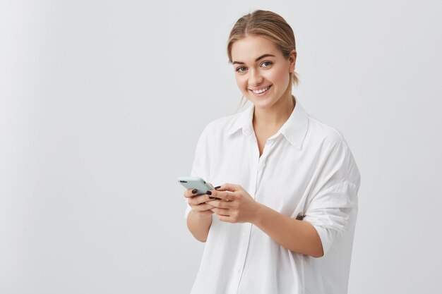 Adorable hipster mujer caucásica con cabello rubio revisando sus noticias o mensajes a través de las redes sociales, usando wi-fi gratis en el teléfono móvil, sonriendo, posando