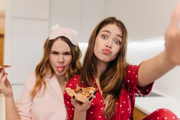 Adorable joven comiendo pizza y jugando. Hermanas positivas divirtiéndose y disfrutando de la comida rápida favorita.