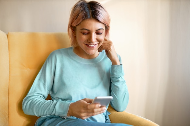 Adorable joven europea con anillo en la nariz y cabello rosado sentada en un sillón con amigos móviles, mensajes de texto, disfrutando de la comunicación en línea.