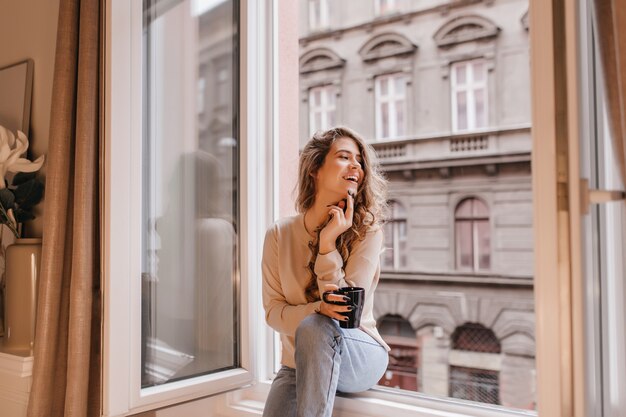 Adorable joven en jeans de moda disfrutando de tiempo libre en casa con una taza de chocolate caliente