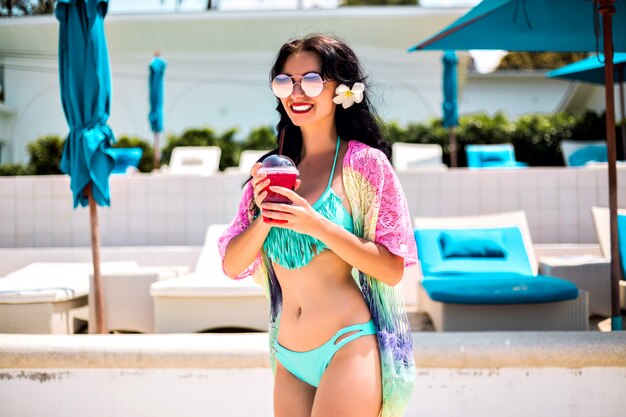 Adorable joven morena sexy posando en el resort de playa de la isla tropical, disfruta de sus vacaciones en el hotel, día caluroso de verano, bebiendo limonada dulce y sonriendo, bikini azul y accesorios, ropa de playa de moda