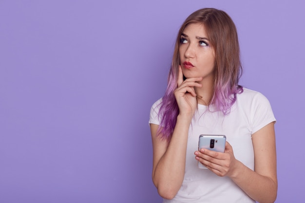 Adorable joven mujer pensativa sosteniendo el teléfono inteligente en las manos, mirando a otro lado con expresión pensativa, manteniendo el dedo en la mejilla, piensa, copie el espacio, posando aislado sobre la pared púrpura.