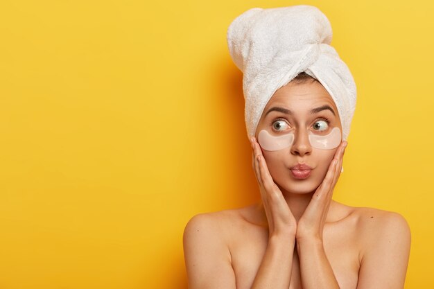 Adorable joven con piel fresca y saludable, toca las mejillas y mira a un lado, usa parches antiarrugas, mantiene los labios redondeados, tiene una toalla en la cabeza, disfruta de un tratamiento de belleza matutino diario aislado en amarillo