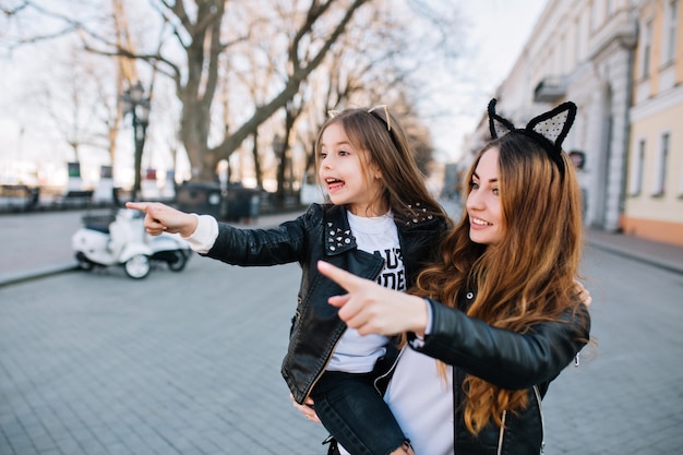 Adorable joven con su hija vio algo interesante al otro lado de la calle. Niña sorprendida en chaqueta de cuero que señala el dedo en el punto de referencia junto a la hermosa mamá.