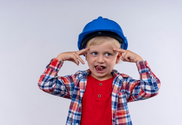 Un adorable y lindo niño con cabello rubio vistiendo camisa a cuadros en casco azul apuntando a su cabeza con los dedos índices