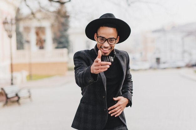 Adorable modelo masculino africano que señala el dedo con expresión de la cara confiada. Retrato al aire libre de hombre negro positivo bien vestido.