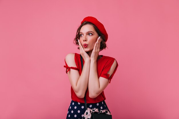 Adorable modelo de mujer caucásica mirando a su alrededor con expresión de cara de sorpresa. Foto interior de bella dama asombrada en boina francesa.