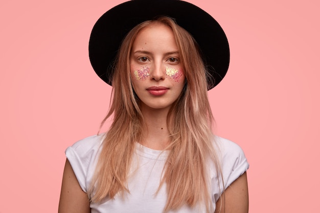 Adorable modelo de mujer joven europea con brillo en la cara, viste un elegante sombrero negro, camiseta blanca, posa sobre una pared rosa, lista para el festival con amigos