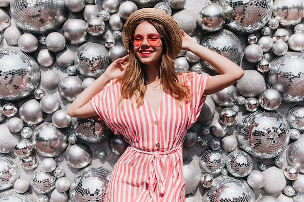 Adorable modelo de mujer con sombrero de paja vintage posando junto a las bolas de discoteca. Mujer rubia relajada en elegante vestido de rayas escalofriante