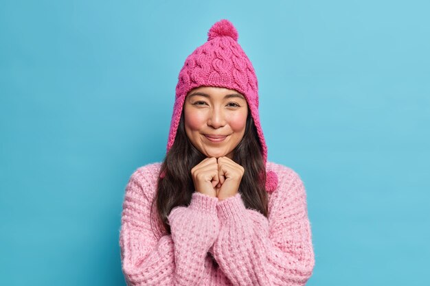 Adorable morena asiática mantiene las manos debajo de la barbilla se ve agradablemente al frente usa gorro de invierno y suéter listo para caminar durante el día frío aislado sobre una pared azul