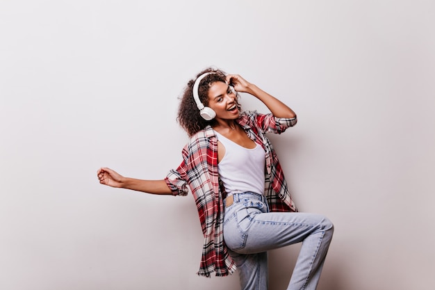 Adorable morena bailando mientras posa en beige. Modelo de mujer africana lleva auriculares y camisa roja