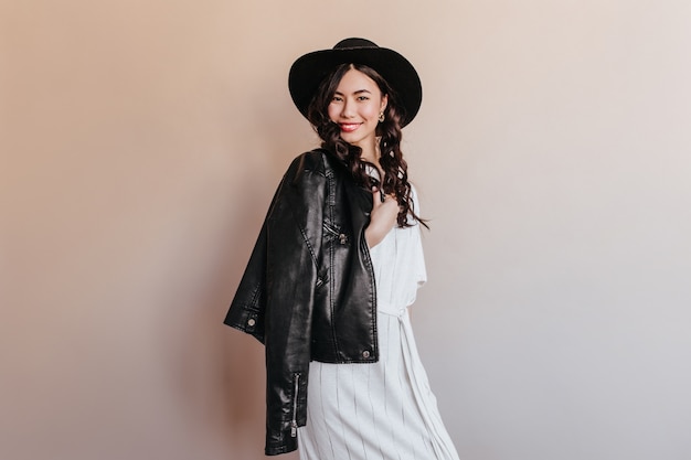 Adorable mujer asiática posando en chaqueta de cuero. Foto de estudio de risa mujer coreana de pie sobre fondo beige.