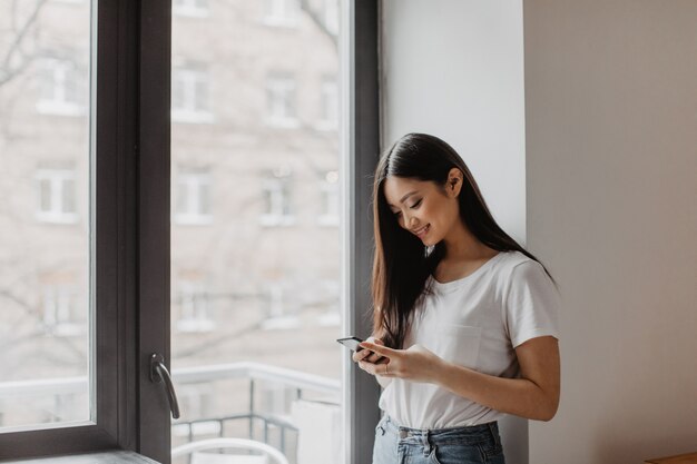 Adorable mujer asiática vestida con top ligero con sonrisa sostiene smartphone y posa junto a la ventana