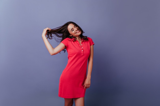 Adorable mujer blanca sonriendo y jugando con su cabello oscuro. Chica debonair en elegante vestido de pie en la pared púrpura.