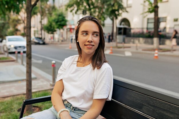 Adorable mujer bonita con cabello oscuro en camiseta blanca está sentada en el banco en la calle de verano en la ciudad en un cálido día de primavera