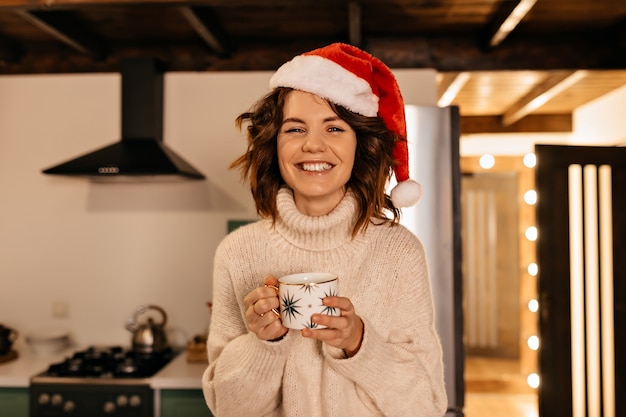 Adorable mujer bonita con pelo rizado con ropa de punto y gorro de Papá Noel sentado en la cocina y esperando la fiesta de Navidad