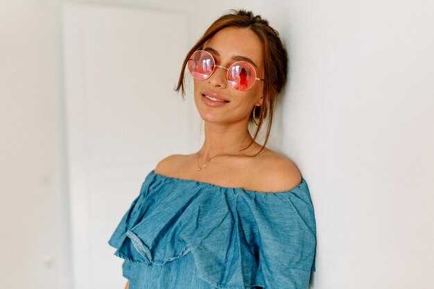 Adorable mujer bronceada con estilo con el pelo oscuro con camiseta azul y gafas redondas rosadas posando en la cámara sobre un fondo aislado