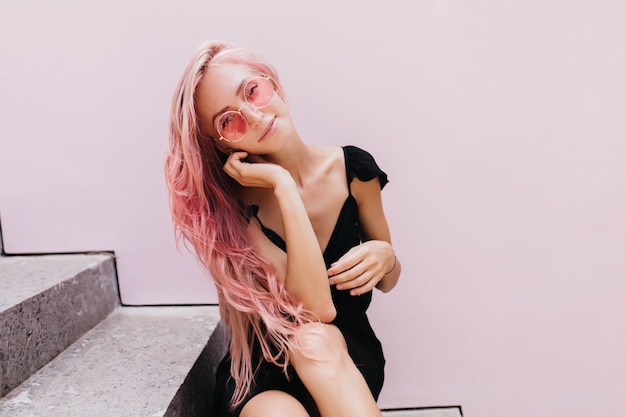 Adorable mujer caucásica con cabello largo rosa descansando sobre las escaleras.