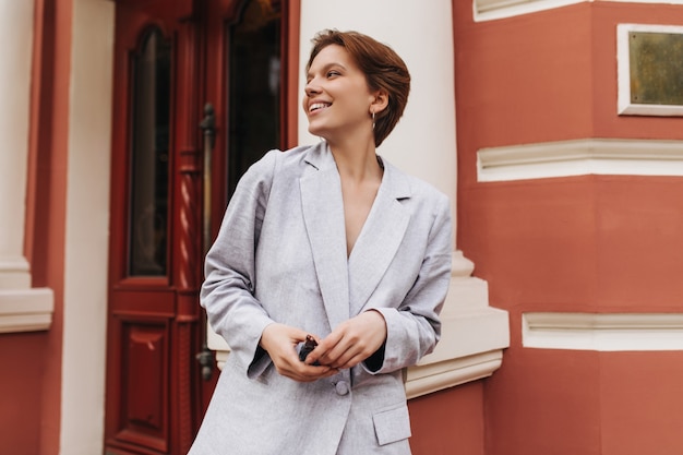 Adorable mujer de chaqueta gris sonriendo y posando afuera. Señora de pelo corto en elegante traje riendo y apoyado en la pared