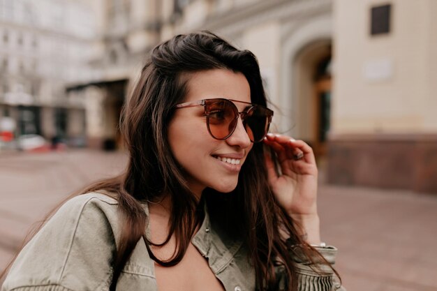 Adorable mujer encantadora con el pelo largo y oscuro con gafas marrones con una sonrisa maravillosa está mirando a un lado