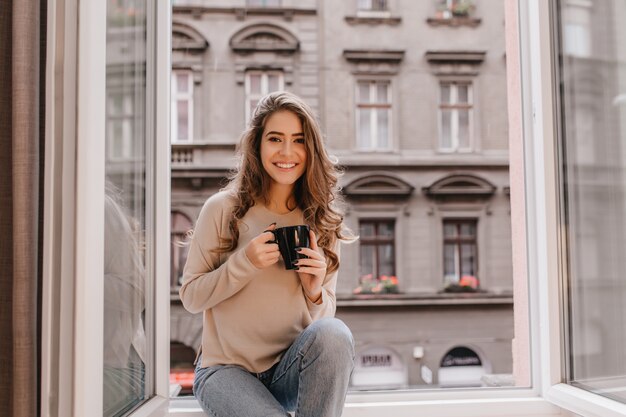 Adorable mujer expresando emociones positivas mientras posa en el alféizar con una taza de café con leche