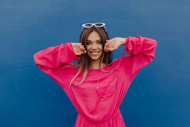 Adorable mujer feliz sonriente con cabello oscuro vestida con camisa rosa posando con las manos levantadas sobre la pared azul aislada