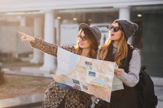 Foto gratuita adorable mujer con gorro de punto gris caminando con un amigo por la ciudad y sosteniendo el mapa. retrato al aire libre de dos encantadoras viajeras mirando algo inetersting en la distancia y señalar con el dedo.