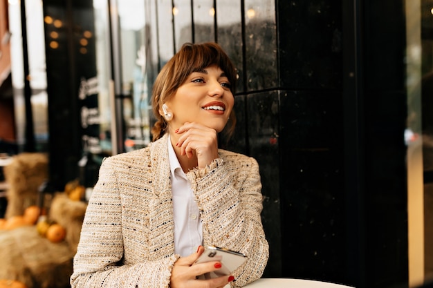 Adorable mujer hermosa en ropa de moda está sentada en un café al aire libre está usando un teléfono inteligente y esperando amigos en el fondo de las luces de la ciudad