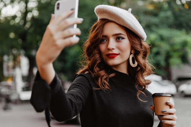 Adorable mujer de jengibre en boina haciendo selfie al aire libre. Señora atractiva tomando café en la calle.