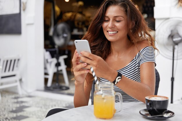 Adorable mujer joven con cabello largo oscuro, vestida con camiseta a rayas en la cafetería, bebe jugo fresco y espresso con el teléfono en las manos