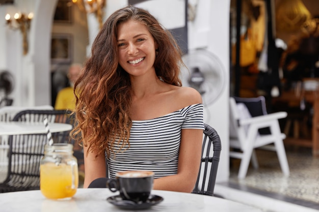 Adorable mujer joven con cabello largo oscuro, vestida con camiseta a rayas en la cafetería, bebe jugo fresco y espresso.