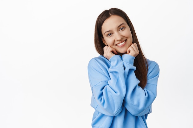 Adorable mujer joven con coqueta sonrisa feliz, inclinar la cabeza, tocar su rostro y sonrojarse, coquetear, mirar y contemplar una hermosa pared blanca.