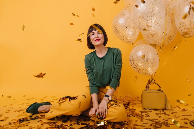 Adorable mujer joven con pelo corto mirando confeti brillante. modelo femenina complacida en ropa de moda sentada junto a globos de helio.