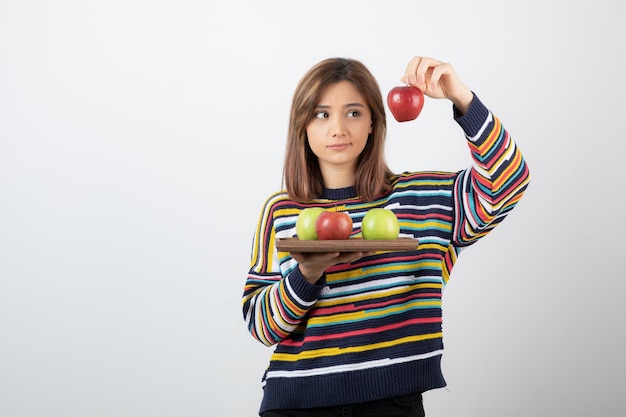 Adorable mujer joven en ropa casual mostrando manzanas rojas sobre pared blanca.