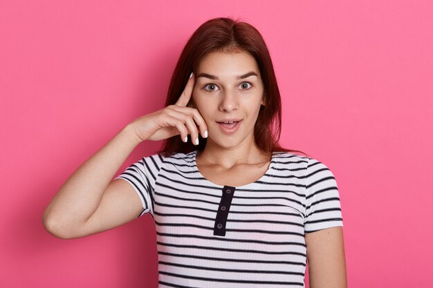 Adorable mujer mantiene los dedos índices en la sien, trata de reunirse con pensamientos, vestida con una camiseta informal a rayas, aislada sobre una pared rosa, tiene una idea, se ve emocionada.