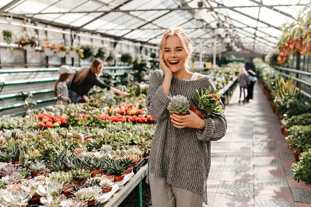 Adorable mujer con moño se ríe sinceramente y posa con variedades de cactus y suculentas.