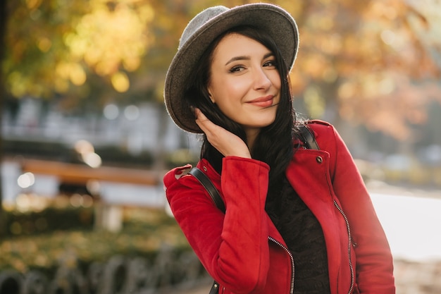 Adorable mujer morena con elegante sombrero gris posando en la pared de naturaleza borrosa