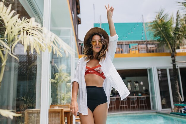 Adorable mujer morena que expresa felicidad mientras posa junto a la piscina del hotel. Retrato al aire libre de moda joven con sombrero y camisa blanca divirtiéndose en el resort.