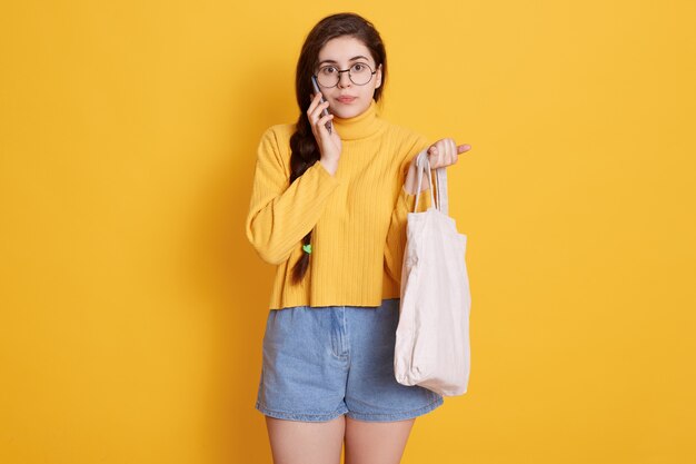 Adorable mujer morena con suéter amarillo y corto, sosteniendo la bolsa en la mano, hablando con su amiga a través de un teléfono inteligente moderno