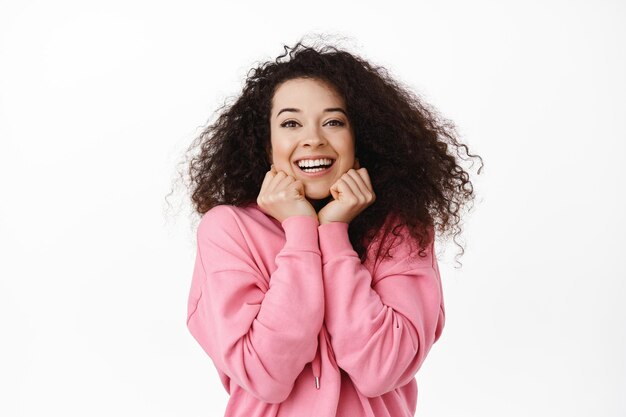 Adorable mujer morena viendo algo hermoso, apoyándose en las manos y sonriendo feliz, mirando con interés y ternura, de pie contra el fondo blanco