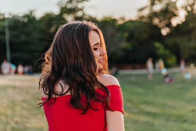 Adorable mujer con peinado elegante posando en el parque. Jocund chica morena caucásica en vestido rojo mirando la hierba.