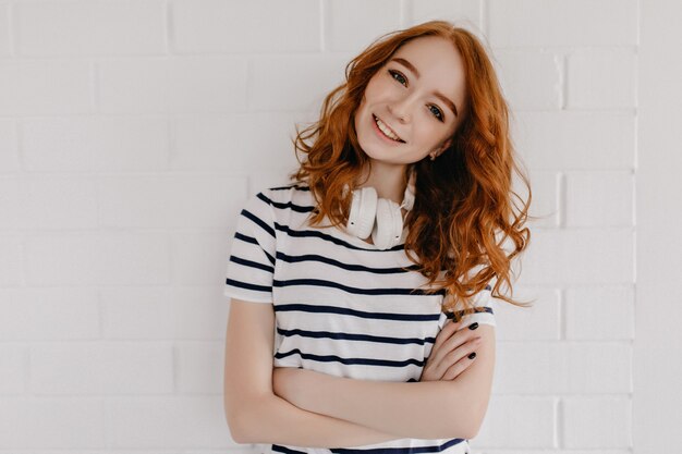 Adorable mujer pelirroja en auriculares de pie con los brazos cruzados. Foto interior de modelo femenino jengibre alegre posando junto a la pared blanca.