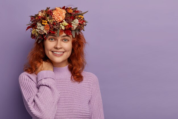 Adorable mujer pelirroja tiene una sonrisa suave, toca el cuello, piel sana, viste una corona otoñal hecha a mano, un jersey morado tejido, se para contra la pared violeta con un espacio vacío.