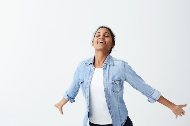 Adorable mujer de piel oscura en camisa vaquera con cabello negro sonriendo con dientes blancos incluso mientras salta. Gente, emociones sinceras, concepto de estilo de vida