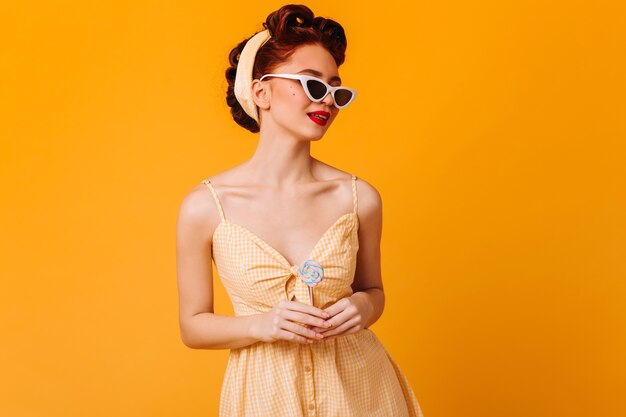 Adorable mujer pinup posando con gafas de sol. Disparo de estudio de niña de jengibre con piruleta aislado en espacio amarillo.