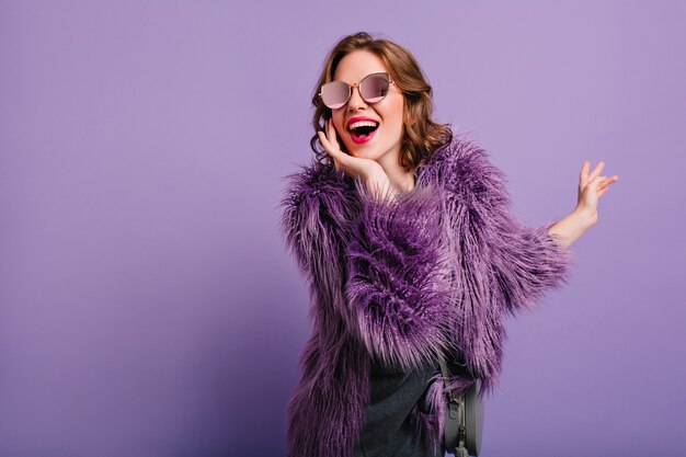 Adorable mujer que expresa verdaderas emociones positivas durante la sesión de fotos en abrigo de piel púrpura