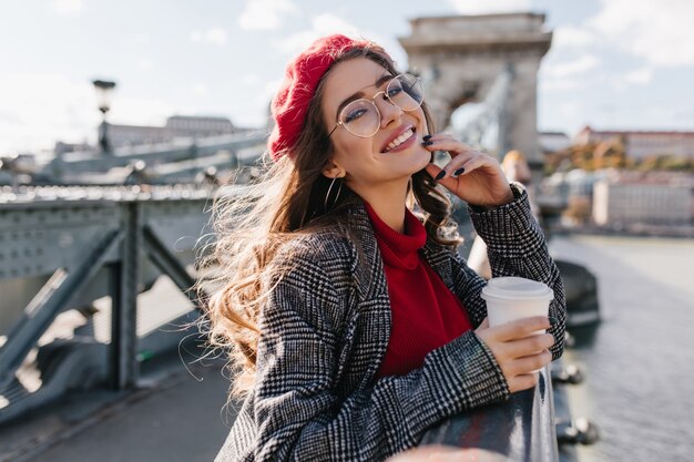 Adorable mujer rizada en lindos vasos tomando café, de pie en el puente con entusiasmo