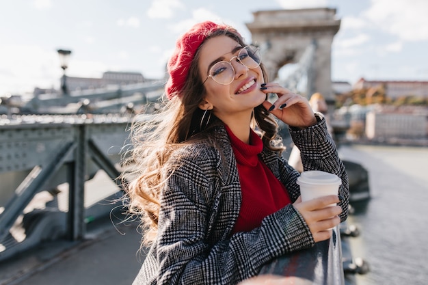 Adorable mujer rizada en lindos vasos tomando café, de pie en el puente con entusiasmo