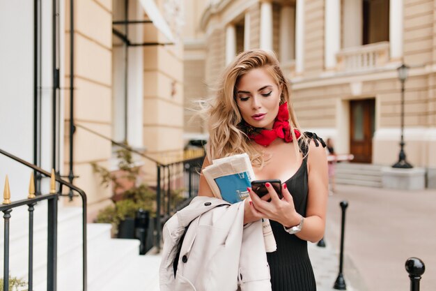 Adorable mujer rubia escribiendo mensaje en el teléfono de pie en medio de la calle